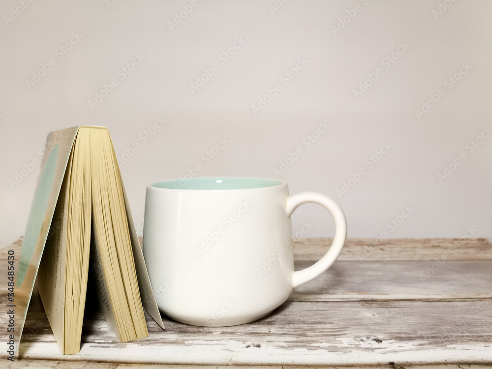 Book and white mug on a wooden table. White background. Space for your text. Image with selective focus and with toning. Book and white mug on a wooden table. White background. Space for your text. Image with selective focus and with toning.