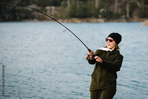 Woman catching a fish, pulling rod while fishing. Girl fishing from pier on lake or pond with text space. Fisherman with rod, spinning reel on river bank. Fishing for pike, perch, carp. Wild nature.