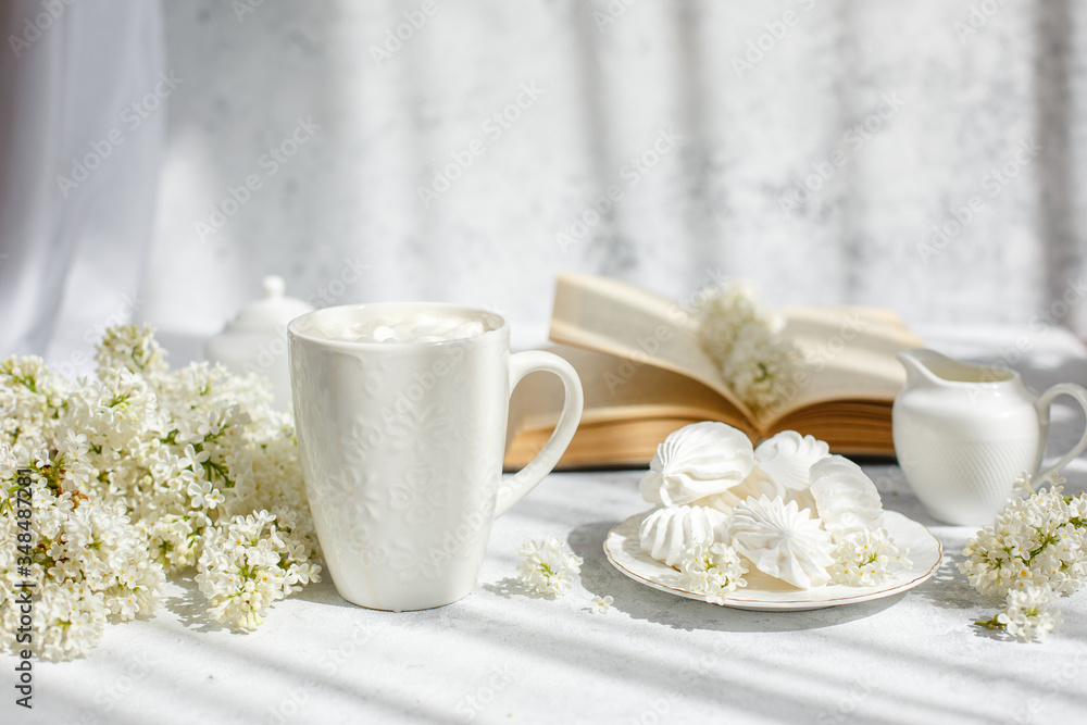 Still life vase with a bouquet of white lilac, a Cup of coffee with marshmallows, a plate of meringue, a sugar bowl and a milk jug , an old book. Postcard good morning.