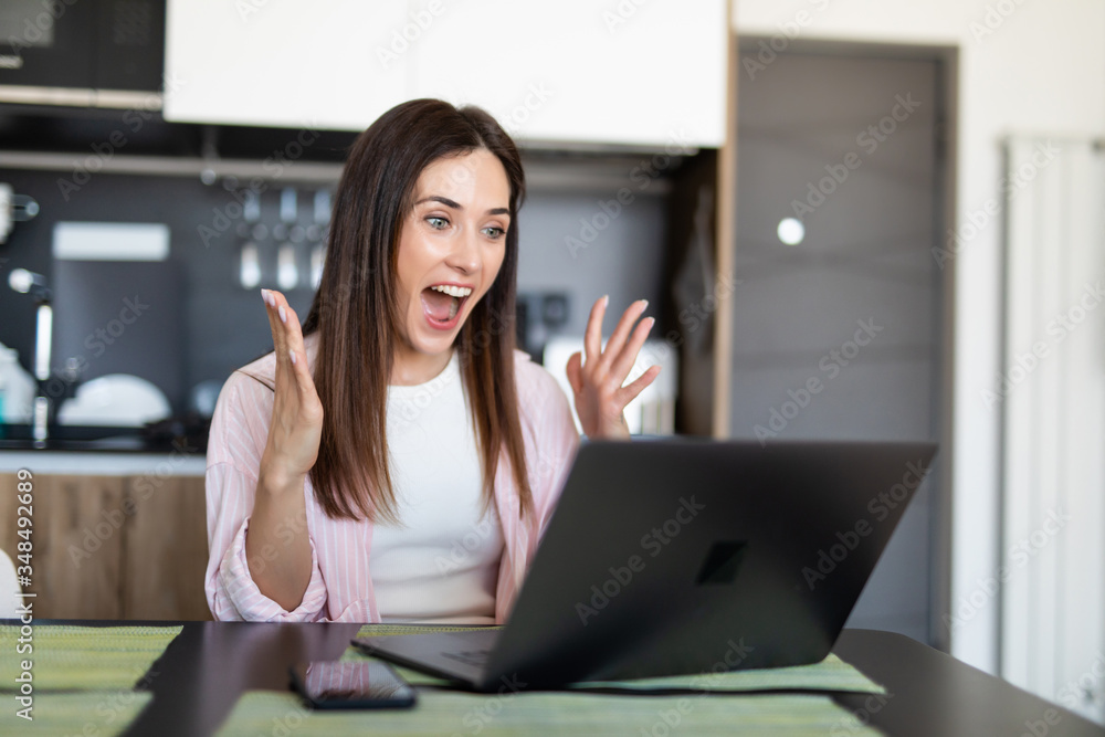 Fototapeta premium Joyful surprised woman putting hands to face looking at laptop while sitting at table at home