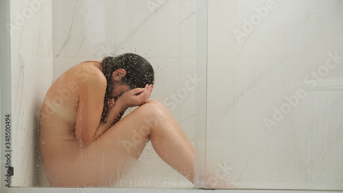 naked lady with long loose wet hair sits on floor behind glass in shower cabin and cries holding hands on face at home