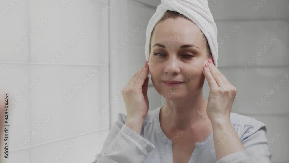 Adult woman with a towel on her head applying lotion and looking in mirror enjoy natural beauty and healthy Adult woman with a towel on her head applying lotion and looking in mirror enjoy natural beauty and healthy