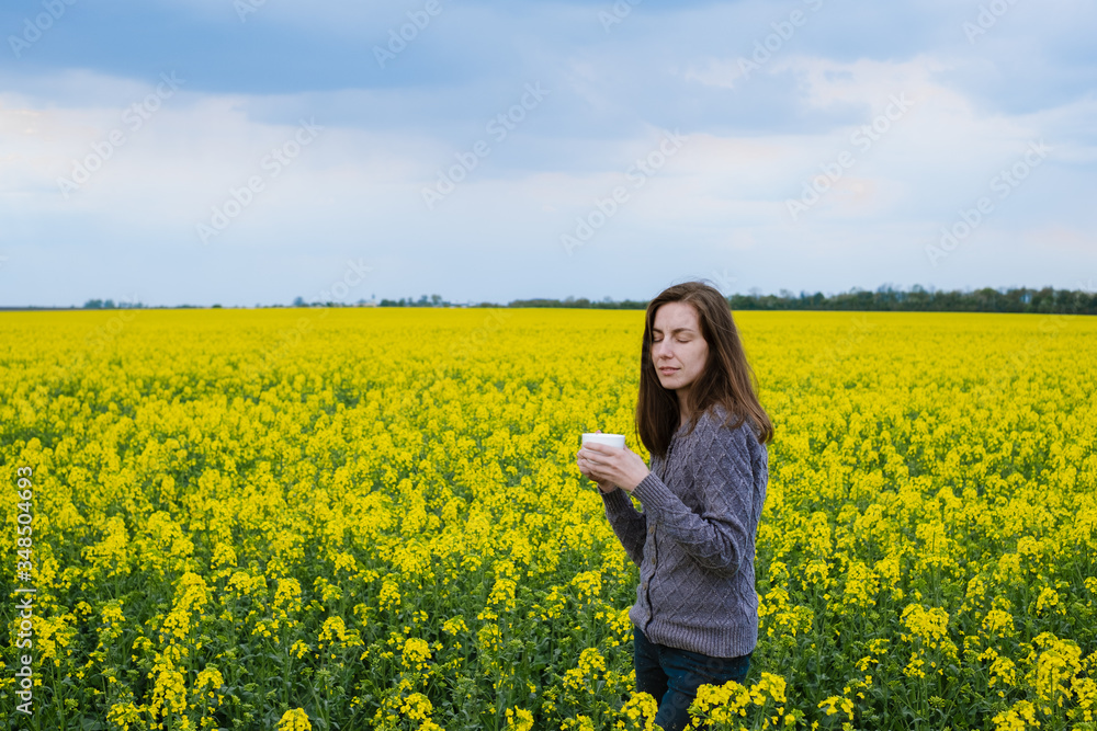 The girl stands in the middle of a flowering rapeseed field and holds a cup of coffee in cloudy weather
The girl stands in the middle of a flowering rapeseed field and holds a cup of coffee in cloudy weather