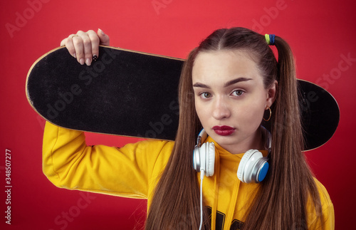 Modern young female in yellow hoodie with headphones on neck holding skateboard on shoulder and looking at camera against red background