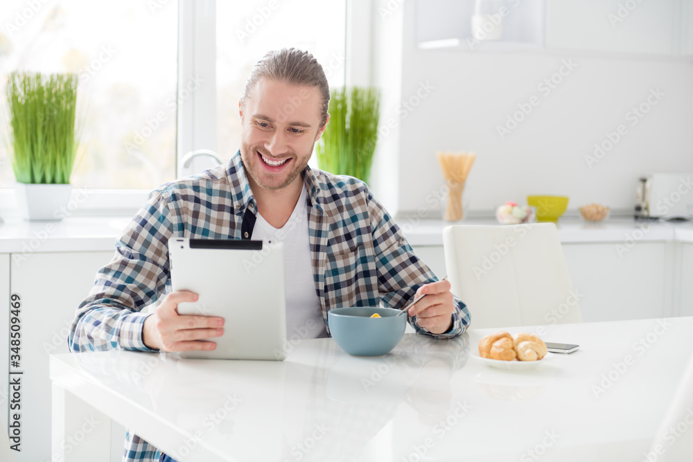 Portrait of his he nice attractive cheerful cheery glad focused guy holding in hand tablet watching video film blog vlog eating snack lunch alone in modern light white interior style kitchen Portrait of his he nice attractive cheerful cheery glad focused guy holding in hand tablet watching video film blog vlog eating snack lunch alone in modern light white interior style kitchen