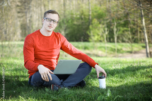 Young man in a red pullover with coffee cup and laptop working outside in a park. Remote work. Digital.