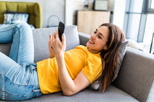 Attractive woman smiling as she reads an sms message on her mobile while relaxing on a sofa in her living room photo