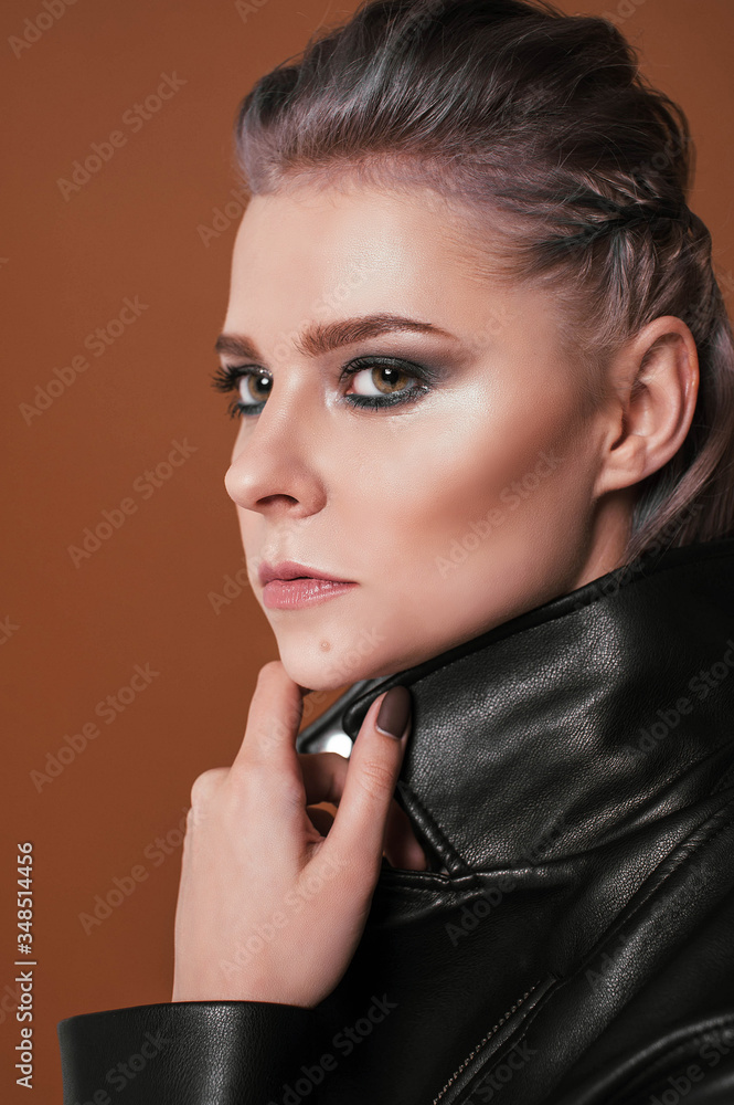 a blonde girl with short hair in black leggings and a leather jacket and boots with a chain and lock around her neck on a cocoa brown background poses a blonde girl with short hair in black leggings and a leather jacket and boots with a chain and lock around her neck on a cocoa brown background poses