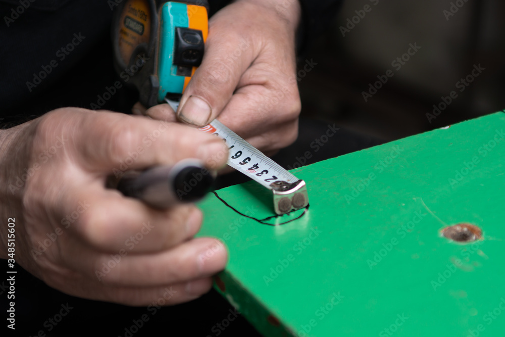 Fototapeta premium An old man in his workshop is making measurements with measuring tape and making marks with black pen on a peace of green woden plate