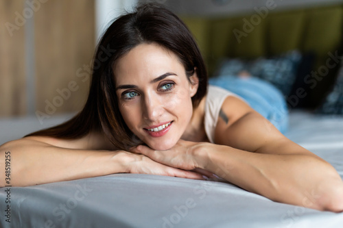 Young woman lying at the end of the bed underneath the quilt and smiling, with her head resting upon her hand with the other in her hair.