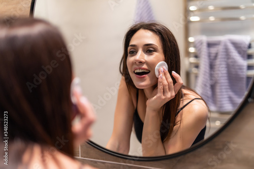 Beautiful young woman is cleaning her face using a cotton disc and smiling while looking in the mirror in the bathroom