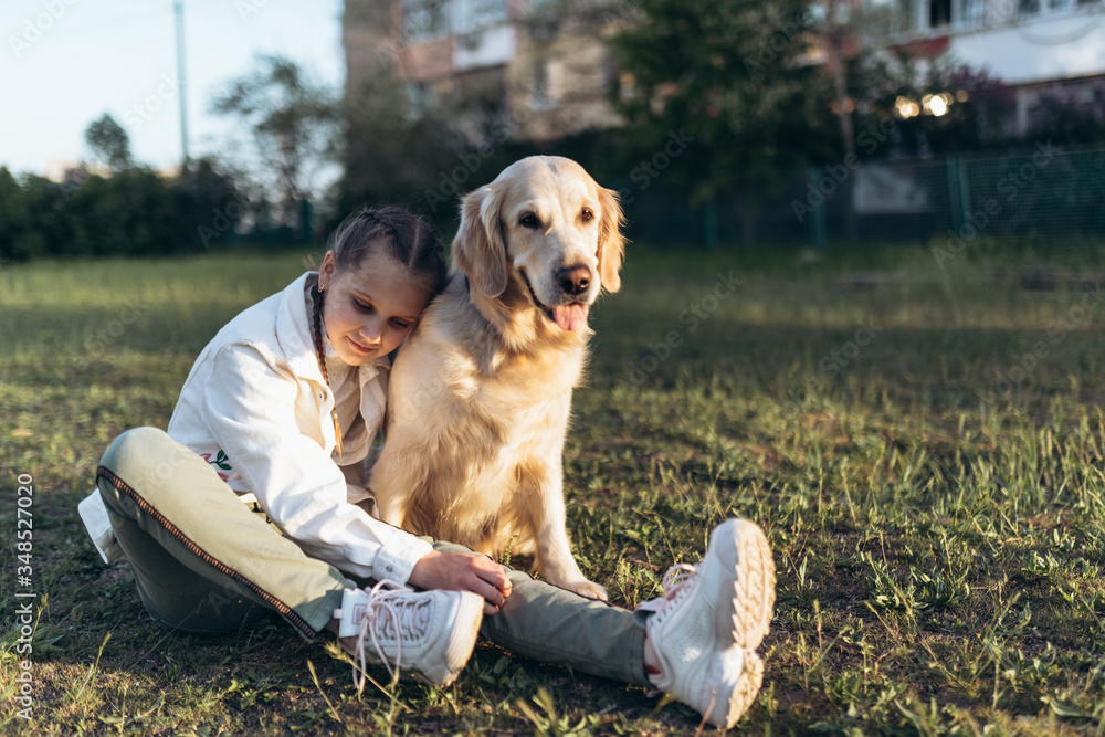 A beautiful girl with two pigtails in white jacket is hugging golden retriever and smiling while sitting on grass. Sunset light.