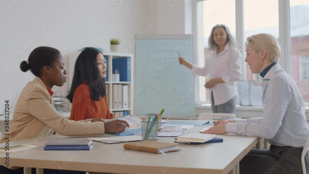 Medium shot of three diverse women sitting at desk in classroom and listening to aged female teacher standing at whiteboard. African woman asking question Medium shot of three diverse women sitting at desk in classroom and listening to aged female teacher standing at whiteboard. African woman asking question