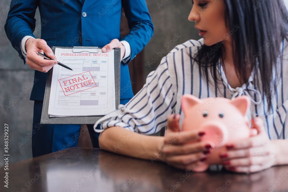 Fototapeta premium Cropped view of woman with piggy bank at table near collector pointing with pen at documents with final notice and foreclosure lettering in room