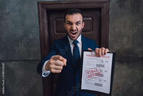 Angry collector with documents with foreclosure lettering pointing with finger and shouting in room