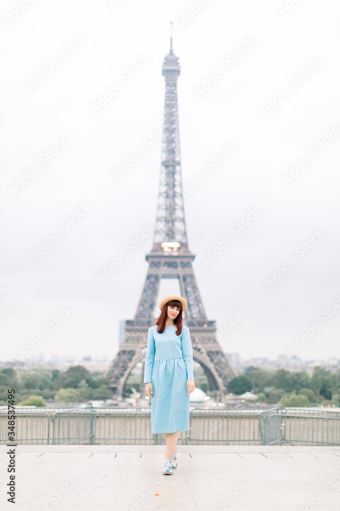 Eiffel Tower in Paris. Cute happy beautiful young Caucasian woman in blue dress and hat, smiling while standing at Trocadero view point in front of Eiffel tower, Paris, France