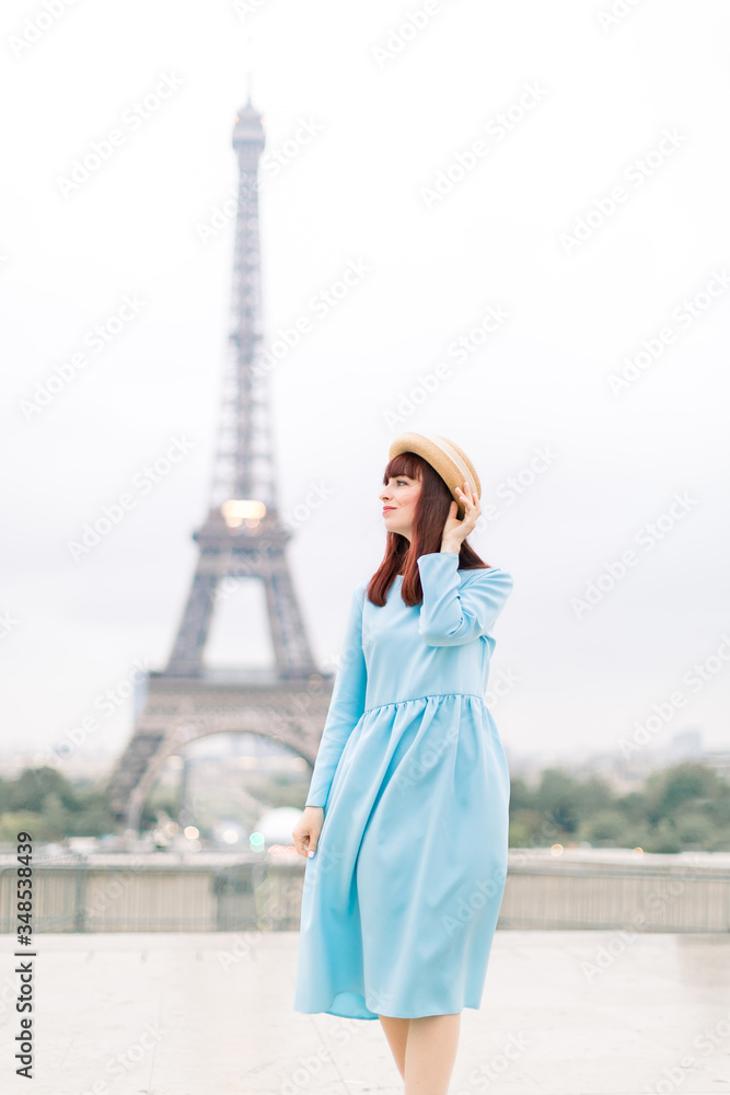 Romantic beautiful girl portrait with tour eiffel on the background. Pretty tender young lady in blue dress and hat touching her red hair and walking in Paris