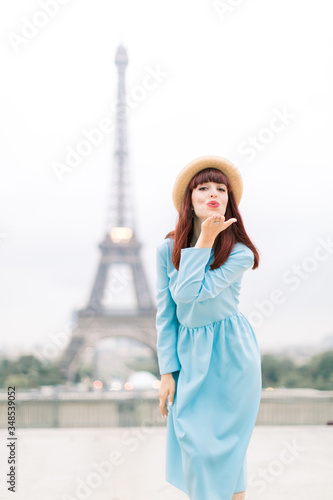 Paris Eiffel Tower. Pretty Caucasian woman in hat and blue dress, smiling happy and making air kiss at Trocadero view point in front of Eiffel Tower in Paris, France