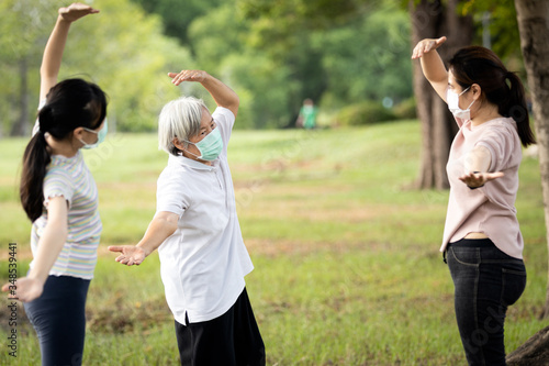 Happy family smiling while exercising at outdoor park,mother,daughter and healthy grandmother are stretching arm workout,wearing surgical mask,enjoy,relaxation after Coronavirus quarantine or Covid-19