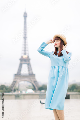 Happy pretty woman wearing hat and blue dress, in Paris with eiffel tower and beautiful morning sky on the background, looking toward and touching her hat, enjoying her vacations © sofiko14