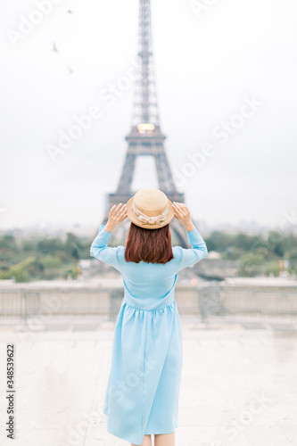 Back view portrait of young attractive woman in hat, blue dress, posing in front of the Eiffel Tower in Paris. Young stylish woman enjoying great view on the Eiffel tower. Travel, tourism concept