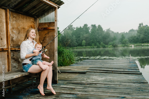 Family fishing holiday. Mother with daughter sitting on wooden pier and catching fish on lake background. Family fishing together on pond. side view. Young mom with child outdoors near river in summer