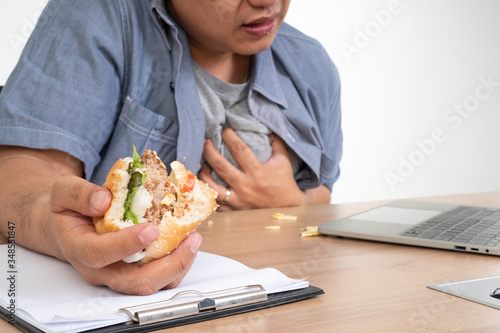 Asian man working and eating a burger on the office desk and heart attack. Concept of a busy businessman cannot work-left balance and not taking care of health eat only junk food