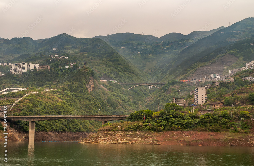Xinling, China - May 6, 2010: Xiling gorge on Yangtze River. 2 road bridges over canyon feeding into Yangtze. Tall mountains in back with sprinkled buildings. Brown sky and green water.