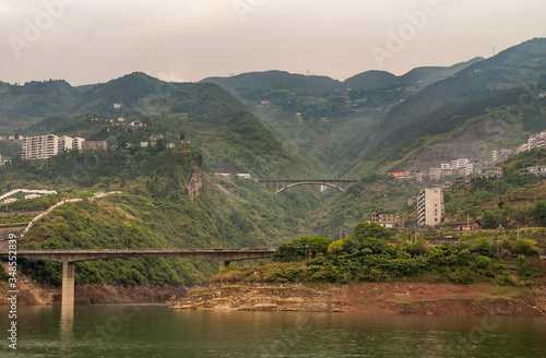 Xinling, China - May 6, 2010: Xiling gorge on Yangtze River. 2 road bridges over canyon feeding into Yangtze. Tall mountains in back with sprinkled buildings. Brown sky and green water. photo