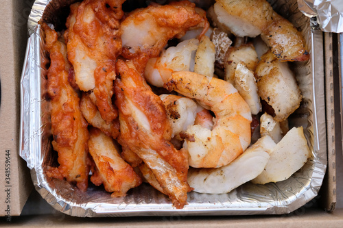 Shrimp and fish slices in an aluminum container. Cod fish in batter and king prawns top view. Home delivery meal with seafood.