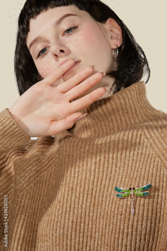 A brunette lady with a blunt bob haircut is covering her mouth. The lady's beige sweater is decorated with an elegant brooch made as a golden dragonfly with malachite enamel and rhinestones. 