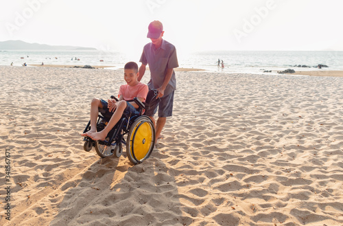 Asian special child and Father in wheelchair on the beach with overflowing light of the sunset, They are happiness in holidays with the travel,Life in the education age and happy disabled kid concept.