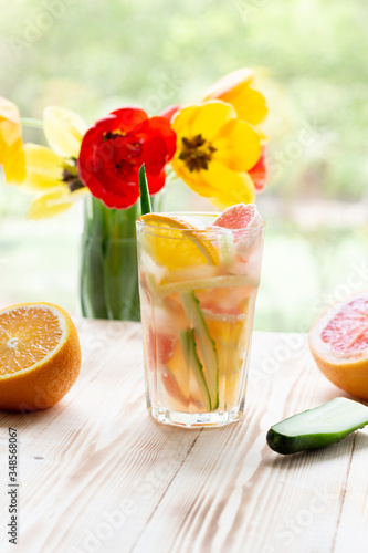 Glass of cocktail with grapefruit, orange, cucumber, ice on wooden background with yellow and red tulips in vase on window