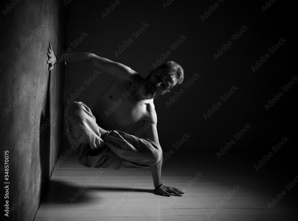 Aged man, naked torso, in brown pants is practicing yoga. Legs in lotus pose, setting one hand against wall, another one on floor