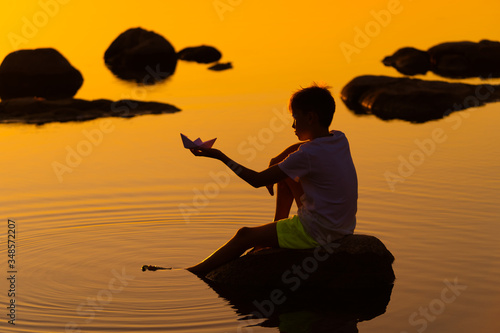 Teen boy with origami ship in hands. Boy sitting on rock near river. Beautiful orange sunset. Origami. River. Lake. Summer vocation