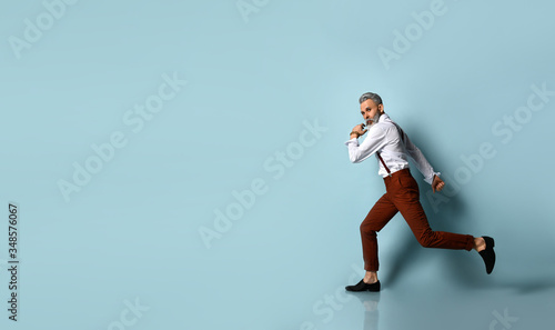 Gray-haired, bearded male in white shirt, brown pants and suspenders, black loafers. He is running against blue background © FAB.1
