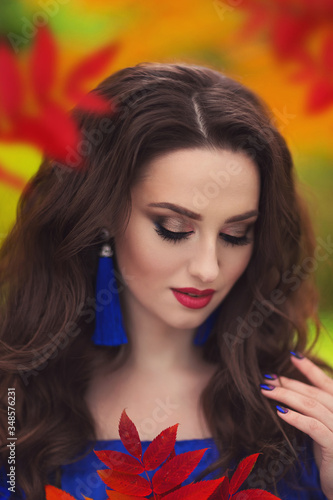 Romantic portrait of a beautiful young girl with blue makeup and red lips, in a long ultramarine blue dress with long blue earrings, on the background of an autumn рark with red leaves