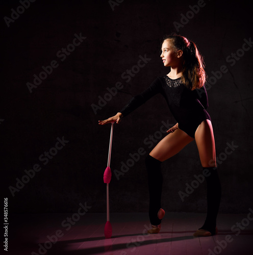 Young smiling girl gymnast in black sport body and uppers standing sideways and holding two pink gymnastic maces