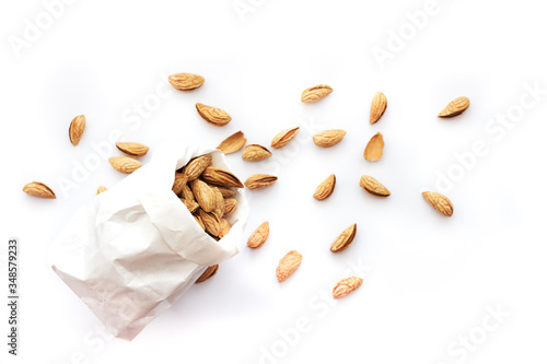 Paper bag stuffed with roasted almonds in peel among almonds scattered on a white background, close up, top view, isolated