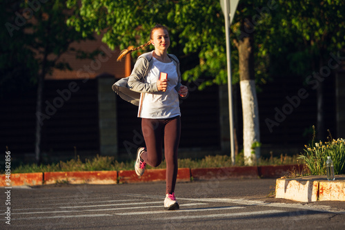 Young female runner, athlete is jogging in the city street in spring sunshine. Beautiful caucasian woman training, listening to music. Concept of sport, healthy lifestyle, movement, activity. © master1305