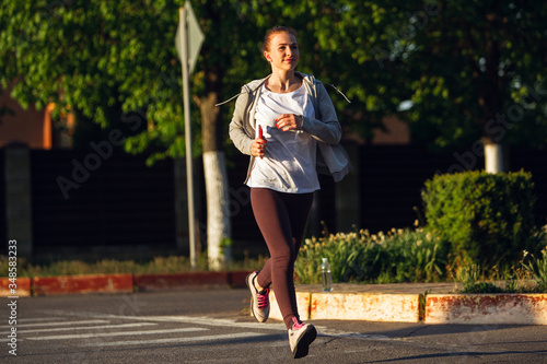 Young female runner, athlete is jogging in the city street in spring sunshine. Beautiful caucasian woman training, listening to music. Concept of sport, healthy lifestyle, movement, activity.