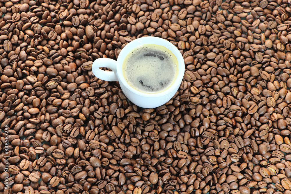 Background from coffee beans, with a white cup in the middle. Roasted coffee beans, randomly laid out on the table. Coffee drink in a white bowl. Background from coffee beans, with a white cup in the middle. Roasted coffee beans, randomly laid out on the table. Coffee drink in a white bowl.