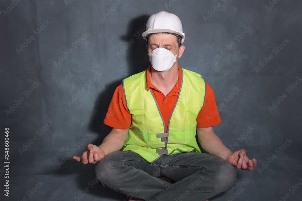 A man in a medical mask sits in a lotus position. Man in a work vest and hard hat. The concept of calm in the workplace during covid-19. A man in a medical mask sits in a lotus position. Man in a work vest and hard hat. The concept of calm in the workplace during covid-19.