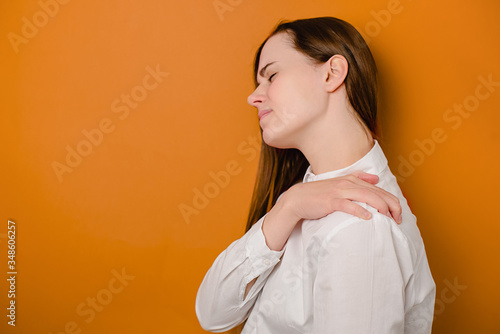Tired unhappy young woman massaging hurt stiff neck isolated on brown studio background, fatigued sad girl rubbing tensed muscles to relieve joint shoulder pain, fibromyalgia concept. Selective focus photo