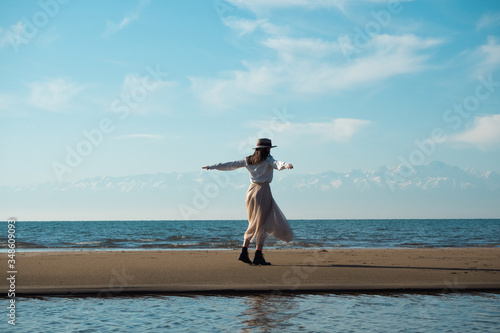 a girl in a white shirt in a hat is spinning on a sandbank on a lake, arms outstretched. A girl stands back to the camera. In the background there is a lake, skyline and mountain peaks in the snow.