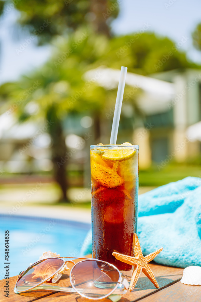 Φωτογραφία Glass of cuba libre cocktail with seashells, sunglasses and blue towel closed to swimming pool
