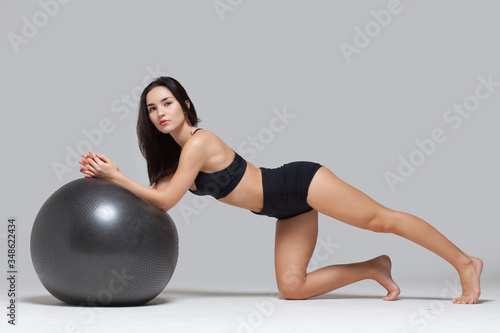 Full length shot of young fitness brunette girl doing exercises with fitness ball looking at camera isolated on grey background