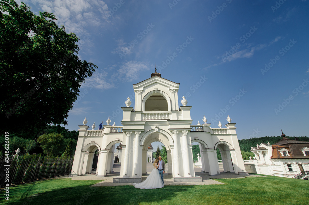 Groom hugs his bride on the green grass against the background of a white arch.