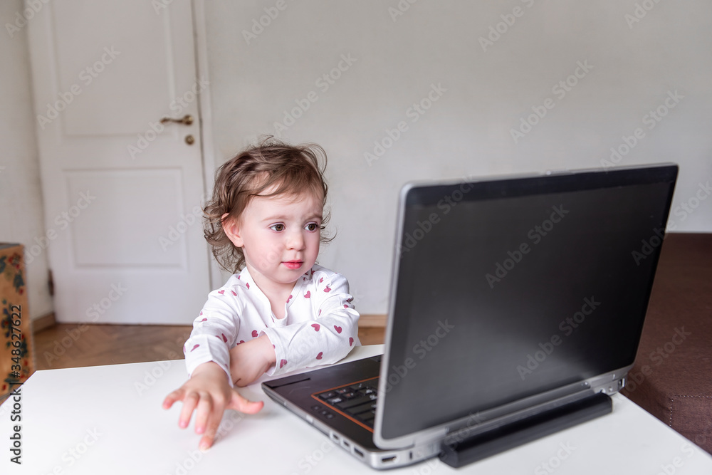 A charming little girl sits behind a laptop and watches and learns from a distance and participates in early development classes. Selective front and back focus, artistic noises. A charming little girl sits behind a laptop and watches and learns from a distance and participates in early development classes. Selective front and back focus, artistic noises.