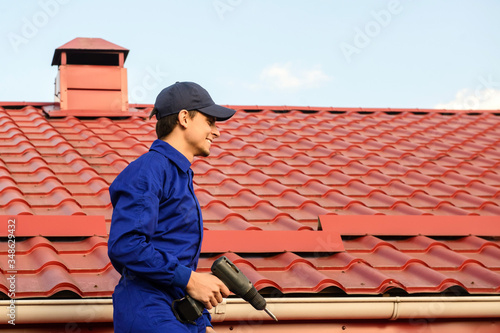 Young happy man contractor worker in blue overalls is repairing a red roof with electric screw driver and have a rest on the roof